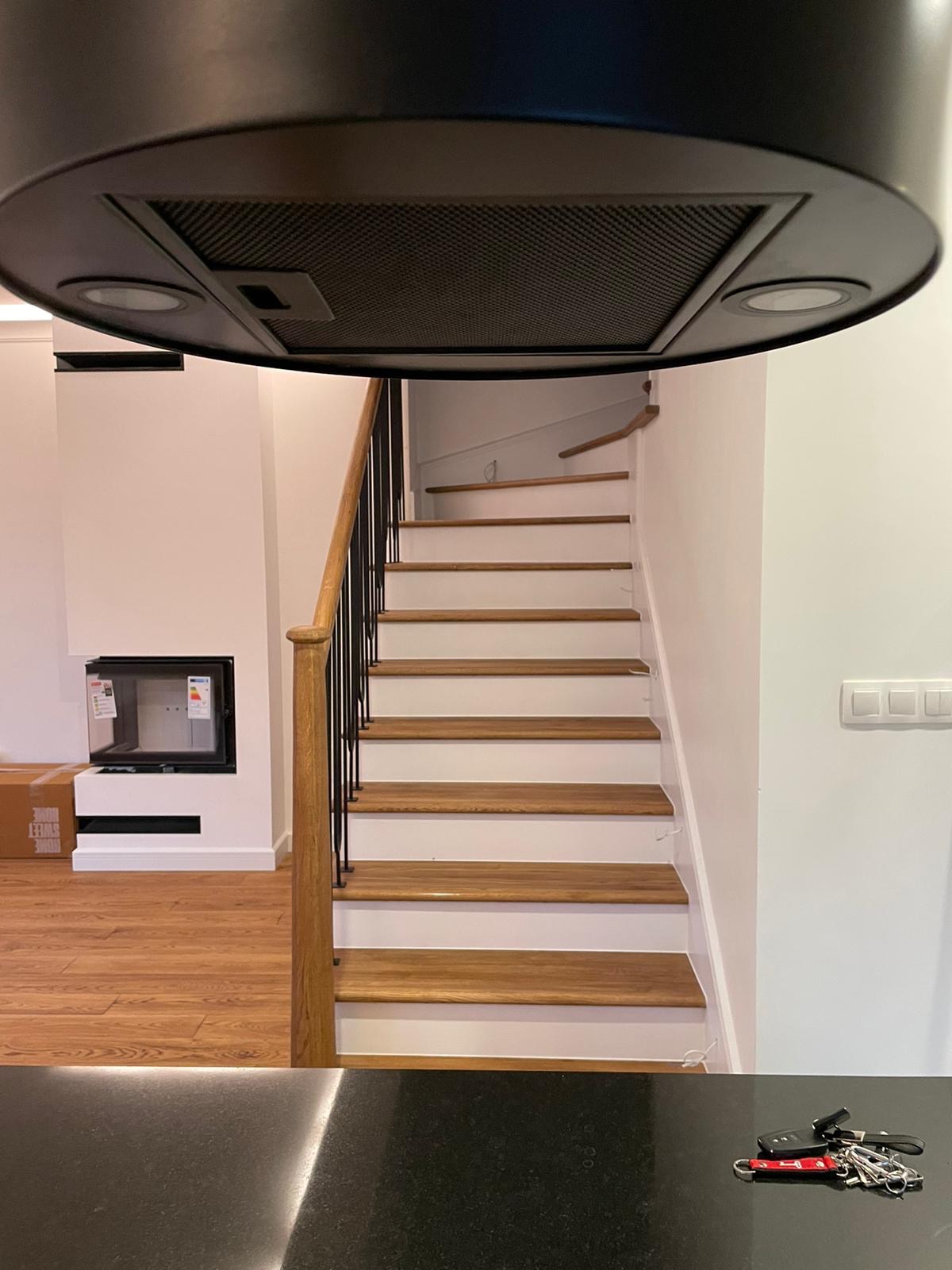 Nowoczesne schody drewniane z czarnymi balustradami i białymi podstopnicami, widoczne z perspektywy blatu kuchennego z kluczami, z fragmentem okapu kuchennego w górnej części kadru i kominkiem w tle.