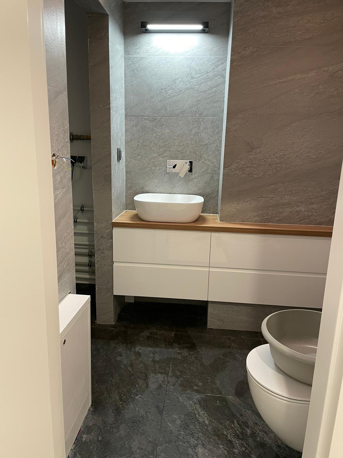 Nowoczesna łazienka z szarymi płytkami imitującymi kamień, białą umywalką nablatową na drewnianym blacie i białymi szafkami pod umywalką, widok zza framugi drzwi.