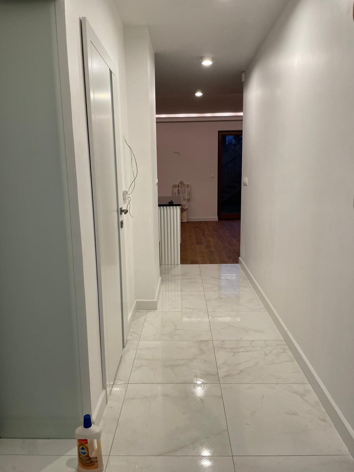 Wykończony korytarz z białymi ścianami, marmurową podłogą i widocznym wejściem do dalszej części domu, z butelką środka czyszczącego w rogu.