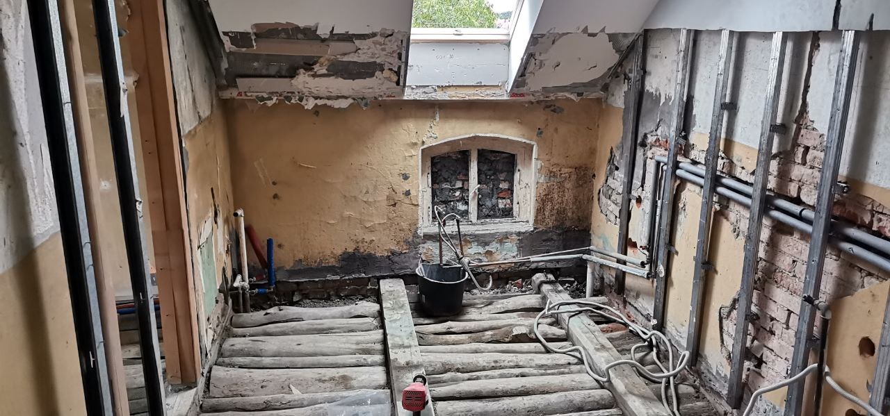 Pokój w trakcie remontu: odsłonięta cegła, metalowe profile do płyt kartonowo-gipsowych, stare okno z kratą, wiadro i kable na drewnianej podłodze.