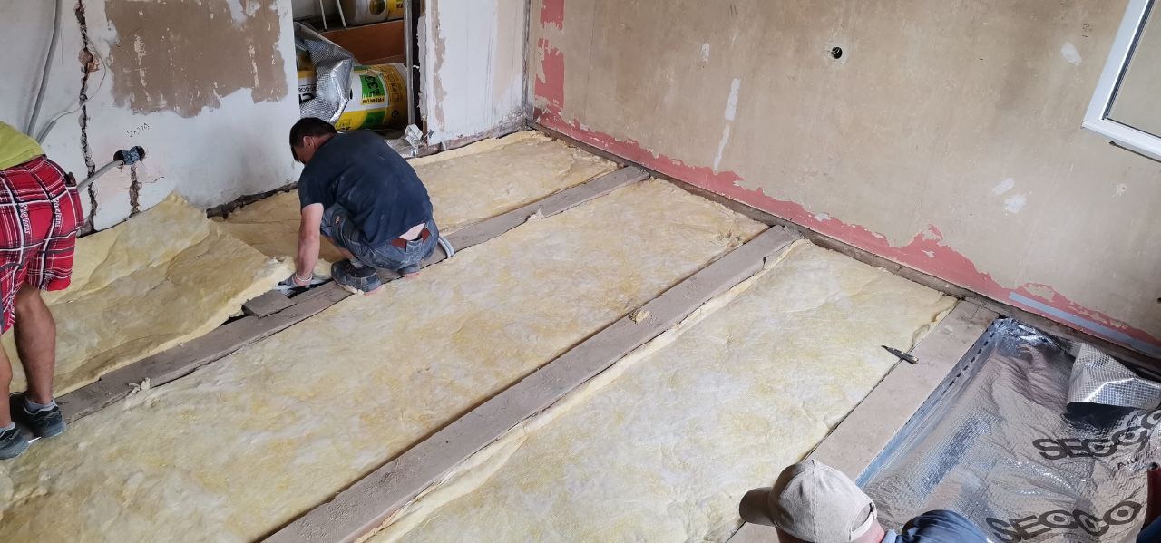 Prace związane z izolacją podłogi wełną mineralną w Szczecinie, widoczne drewniane legary i fragment folii paroizolacyjnej.