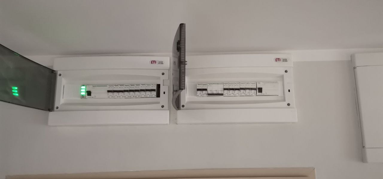 Dwa białe panele elektryczne ETI zamontowane na ścianie, jeden z otwartą osłoną ukazującą włączone zielone diody LED.