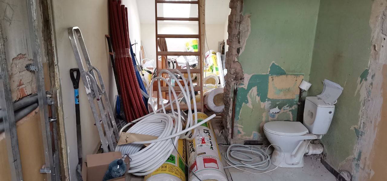 Łazienka w trakcie demontażu: widoczne rury, drabina, materiały izolacyjne oraz częściowo zdemontowana toaleta z odsłoniętymi warstwami ścian.