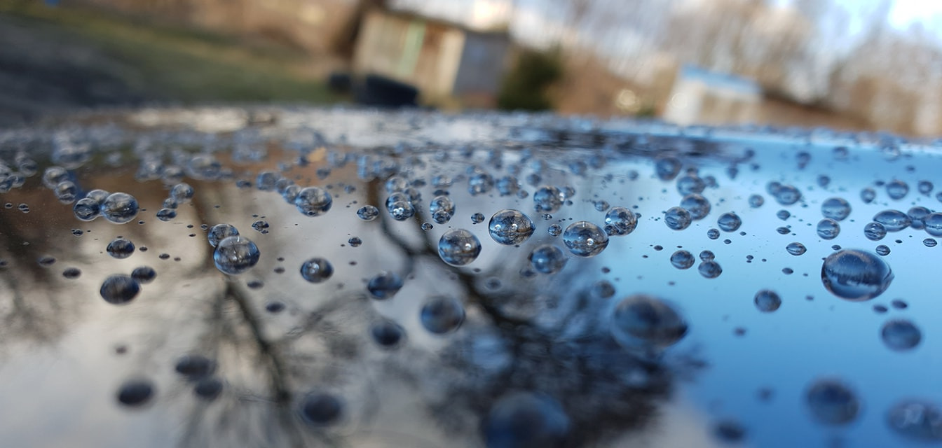 Krople wody na lakierowanej powierzchni samochodu, z odbiciem drzew i nieba w każdej kropli, ukazujące efekt hydrofobowy po woskowaniu.
