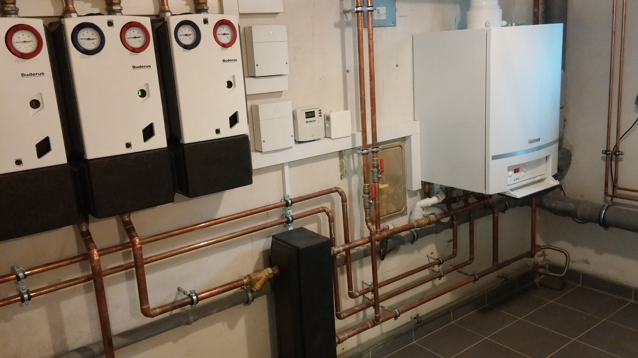 Instalacja grzewcza z kilkoma urządzeniami marki Buderus, z widocznymi rurami miedzianymi i elementami sterującymi na ścianie w kotłowni.