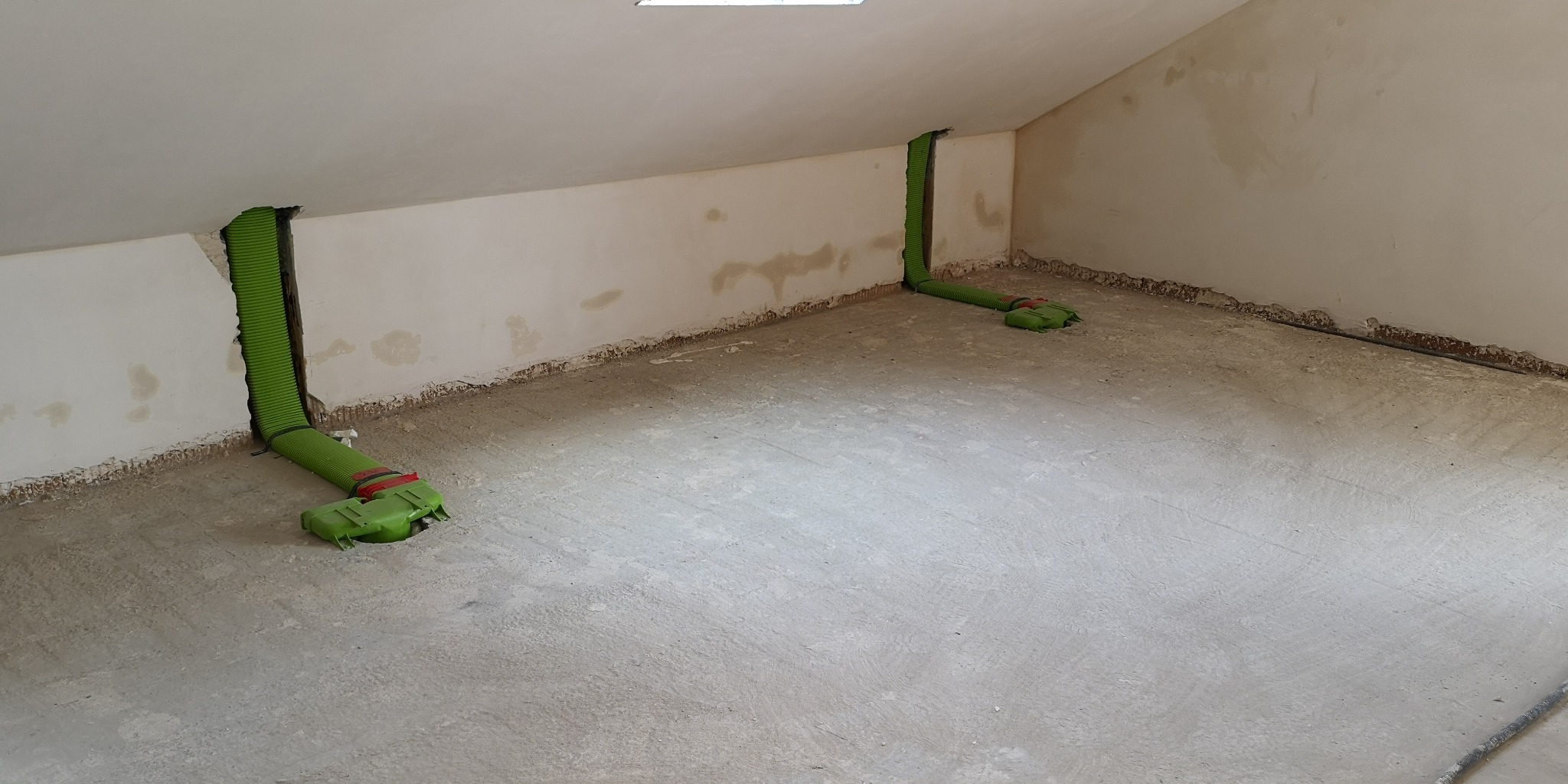 Wnętrze poddasza z dwoma zielonymi, karbowanymi przewodami wentylacyjnymi wystającymi ze ścian, podłączonymi do elementów instalacji w podłodze.