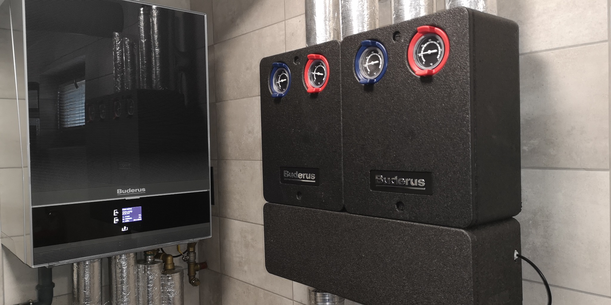 Nowoczesny system grzewczy Buderus z dwoma modułami, z widocznymi manometrami w czerwonych i niebieskich obudowach, zainstalowany na tle jasnych płytek ceramicznych.