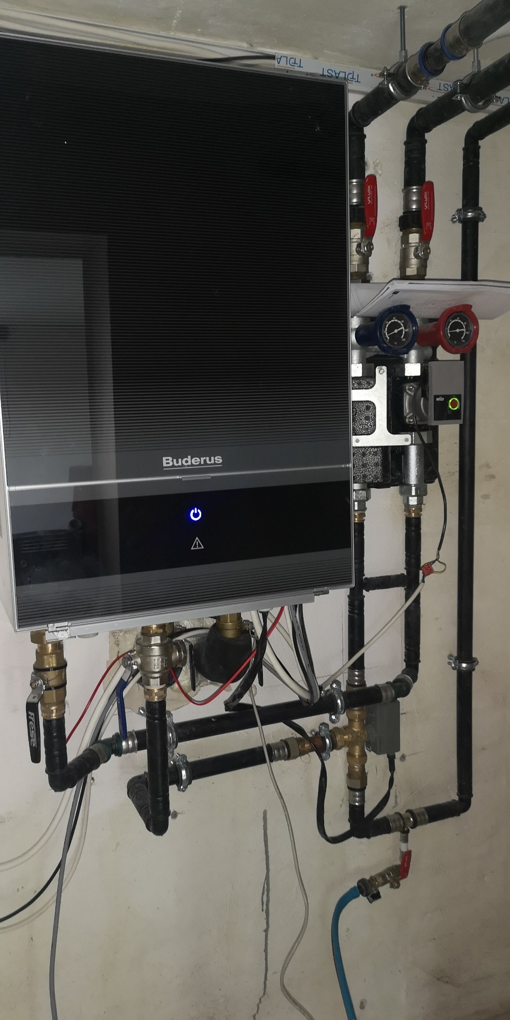 Czarny, wiszący kocioł grzewczy Buderus z podłączonymi rurami i zaworami, zamontowany na jasnej ścianie.