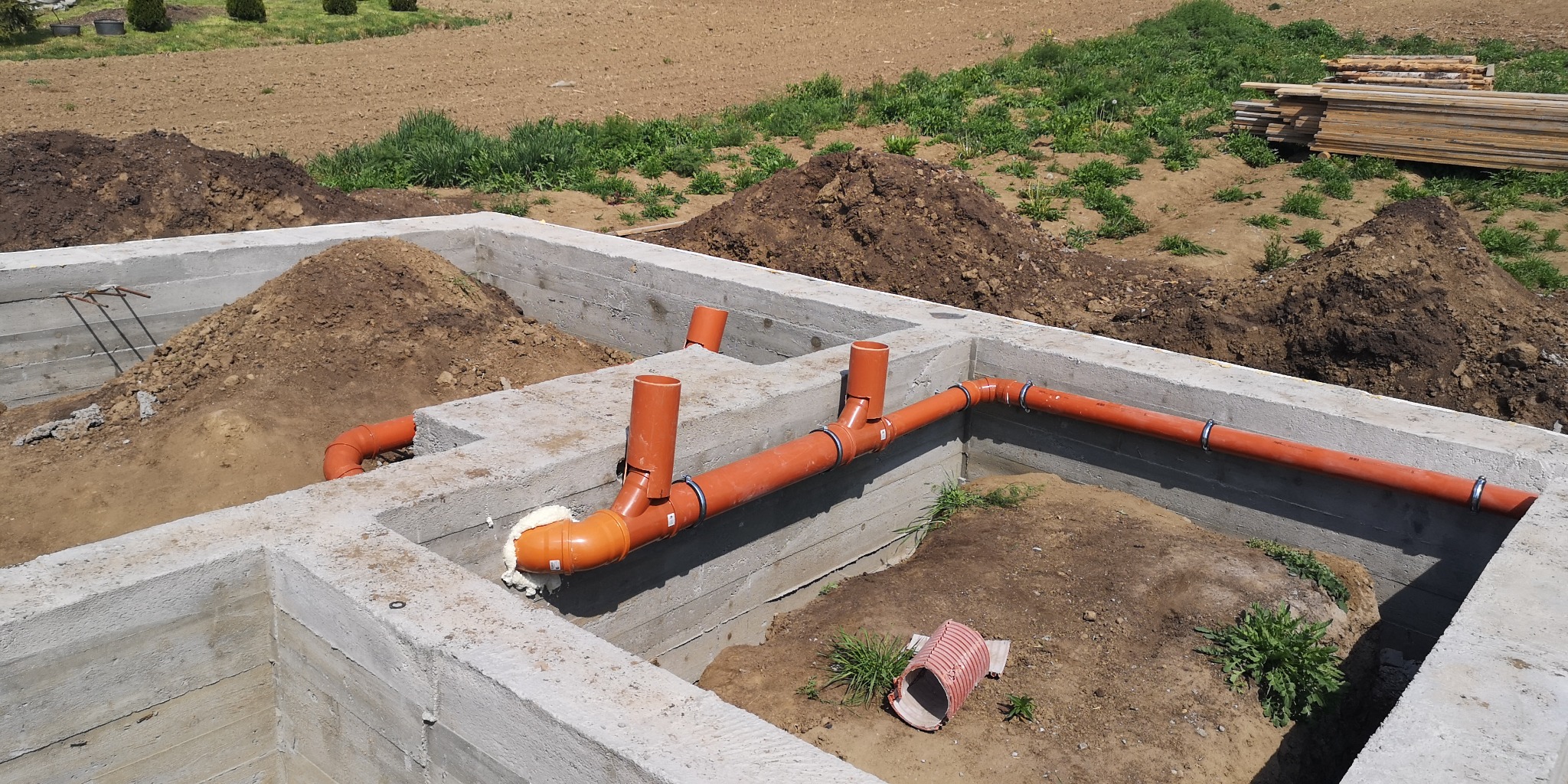 Fundament budynku z betonowych bloków, z zainstalowaną pomarańczową instalacją kanalizacyjną, uszczelnioną pianką montażową, widoczne kupki ziemi i deski w tle.