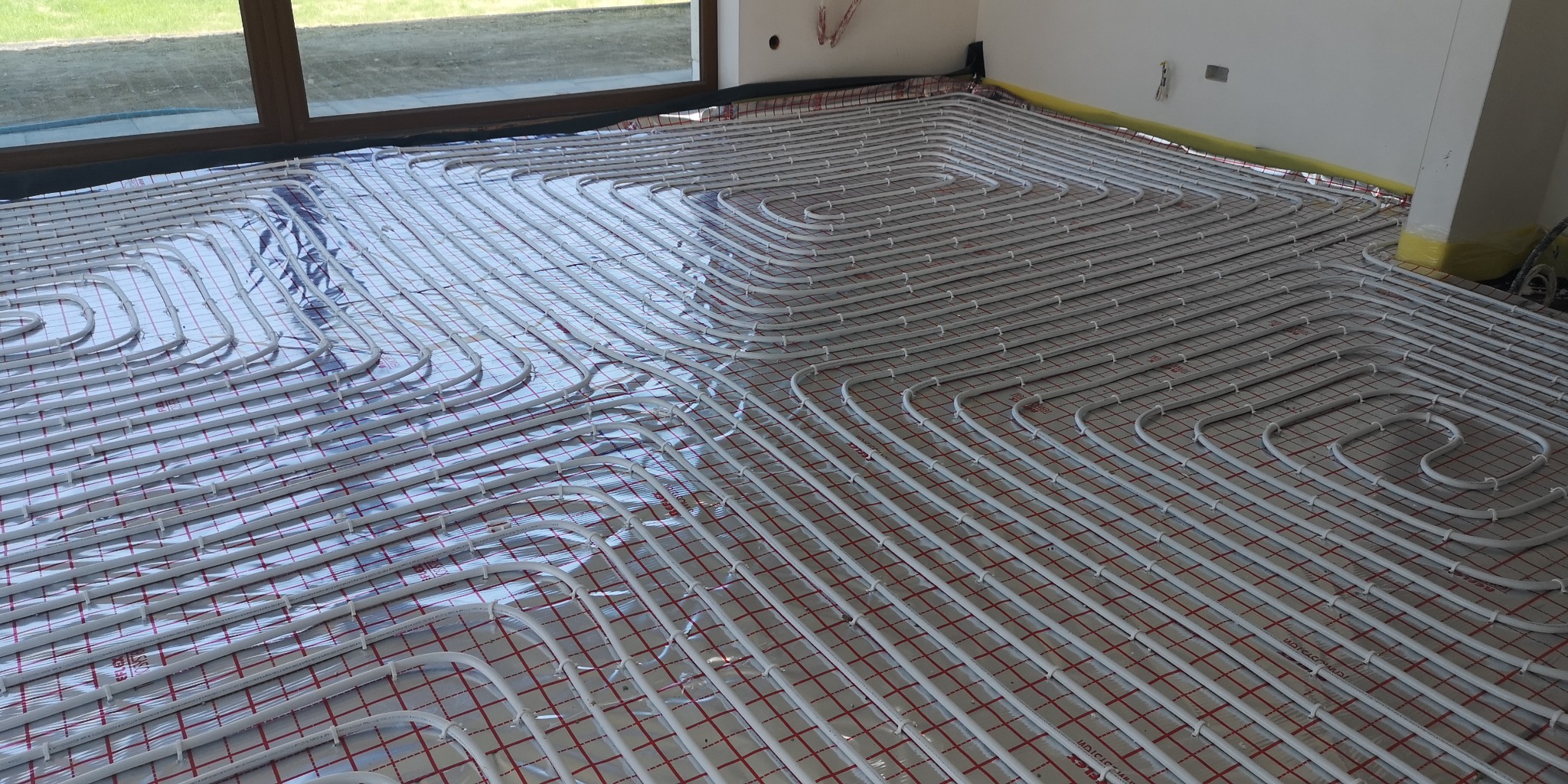 Montaż ogrzewania podłogowego z rur na folii refleksyjnej z siatką, widoczne okno i fragment ściany.