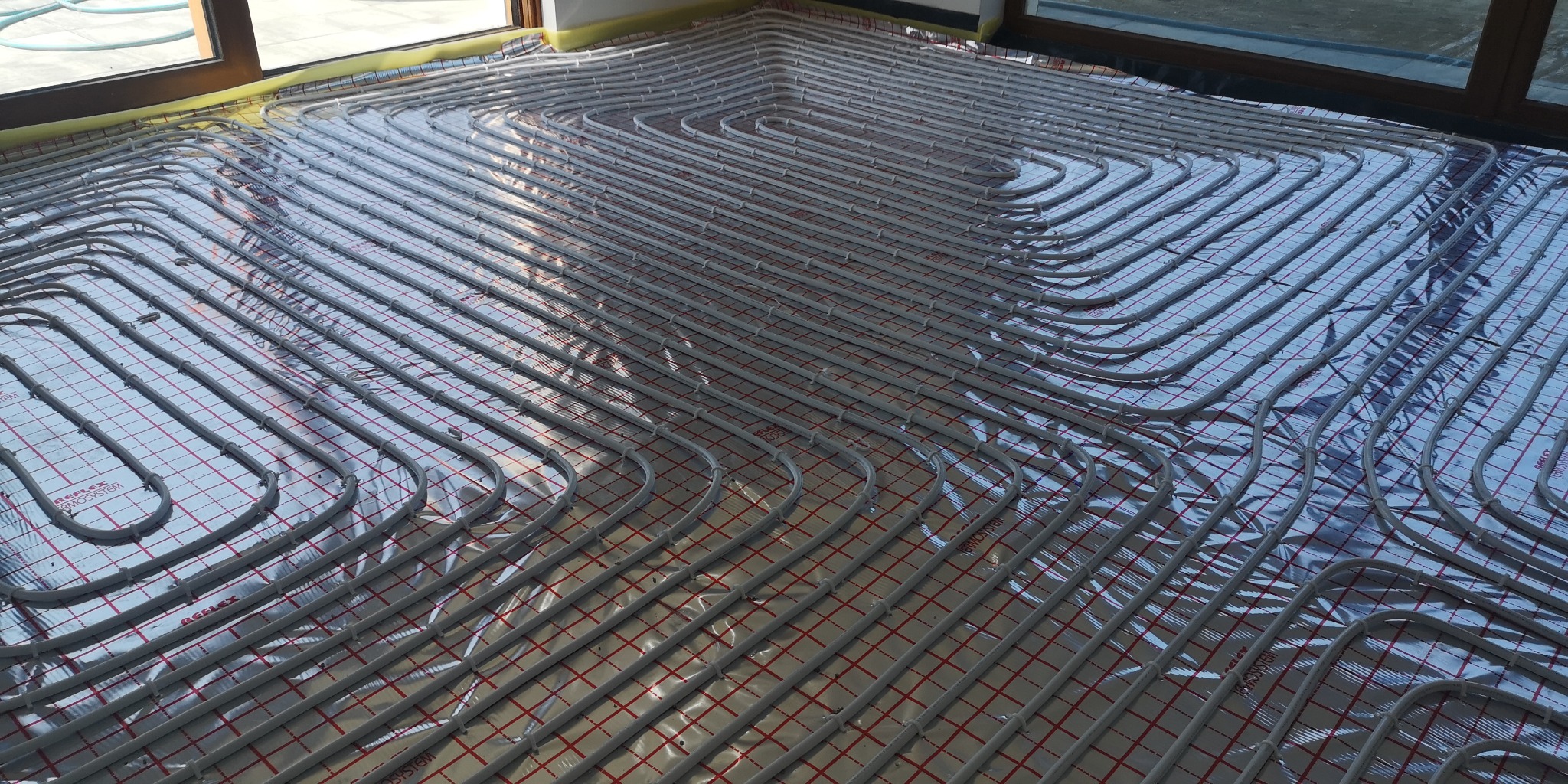 Instalacja ogrzewania podłogowego z rur grzewczych ułożonych w pętle na folii refleksyjnej z czerwoną kratką, widoczne okna balkonowe.