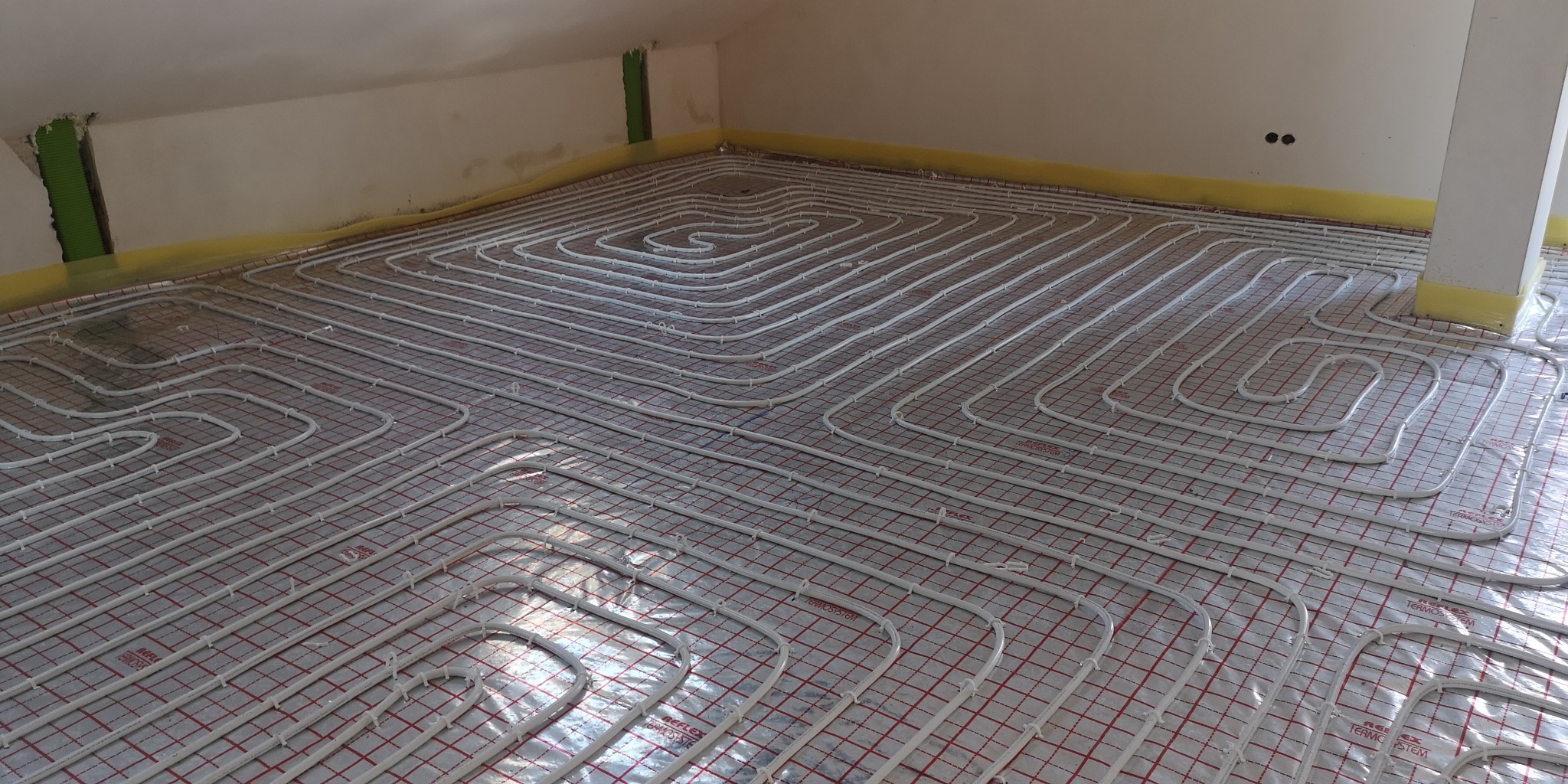 Białe rury ogrzewania podłogowego ułożone spiralnie na czerwonej siatce w surowym wnętrzu z widocznymi niedoskonałościami ścian i przewodami.