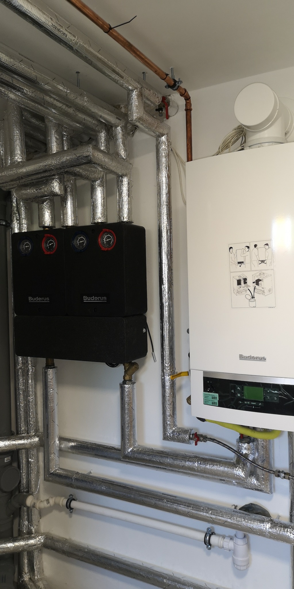 Złożona instalacja grzewcza z dwoma czarnymi modułami i białym kotłem grzewczym marki Buderus, rury izolowane folią aluminiową, widoczne manometry z czerwonymi i niebieskimi wskazówkami.