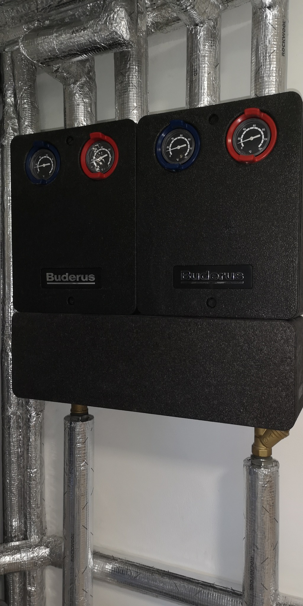 Czarny moduł pompy ciepła Buderus z manometrami w czerwonych i niebieskich obwódkach, podłączony do izolowanych rur z folii aluminiowej.