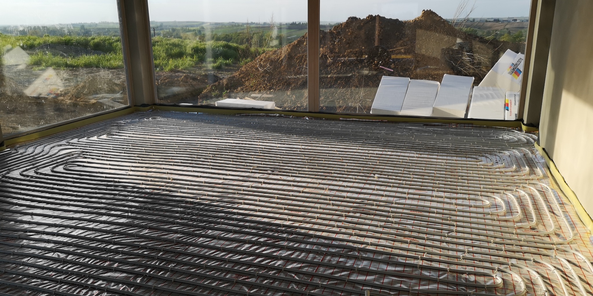 Instalacja ogrzewania podłogowego w nowym budynku z widokiem na krajobraz za oknem i stertę ziemi.