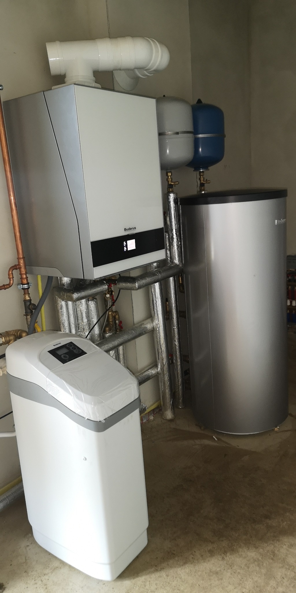 Zamknięte pomieszczenie techniczne z zamontowanym srebrnym zasobnikiem ciepłej wody, białą stacją uzdatniania wody oraz instalacją rur w izolacji.