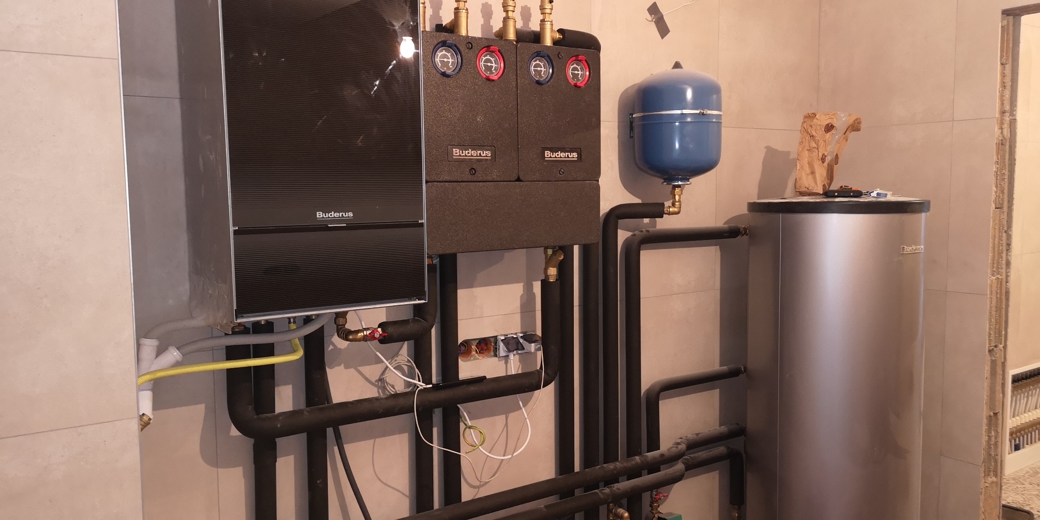 Zainstalowany system grzewczy Buderus z czarnymi rurami, zbiornikiem i elementami kontrolnymi w pomieszczeniu wyłożonym jasnymi płytkami. Na górze zbiornika leżą narzędzia i papierowa torba.