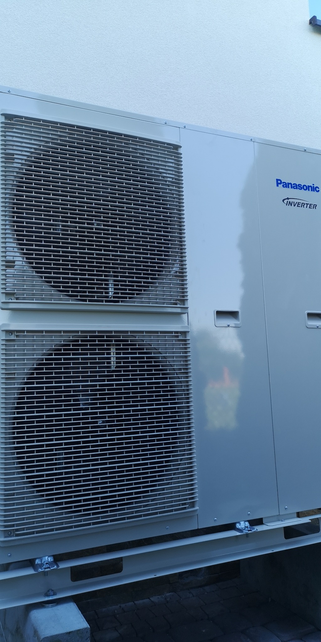 Zewnętrzna jednostka klimatyzacyjna Panasonic Inverter z dwoma wentylatorami, zamontowana na betonowych bloczkach przy białej ścianie budynku.