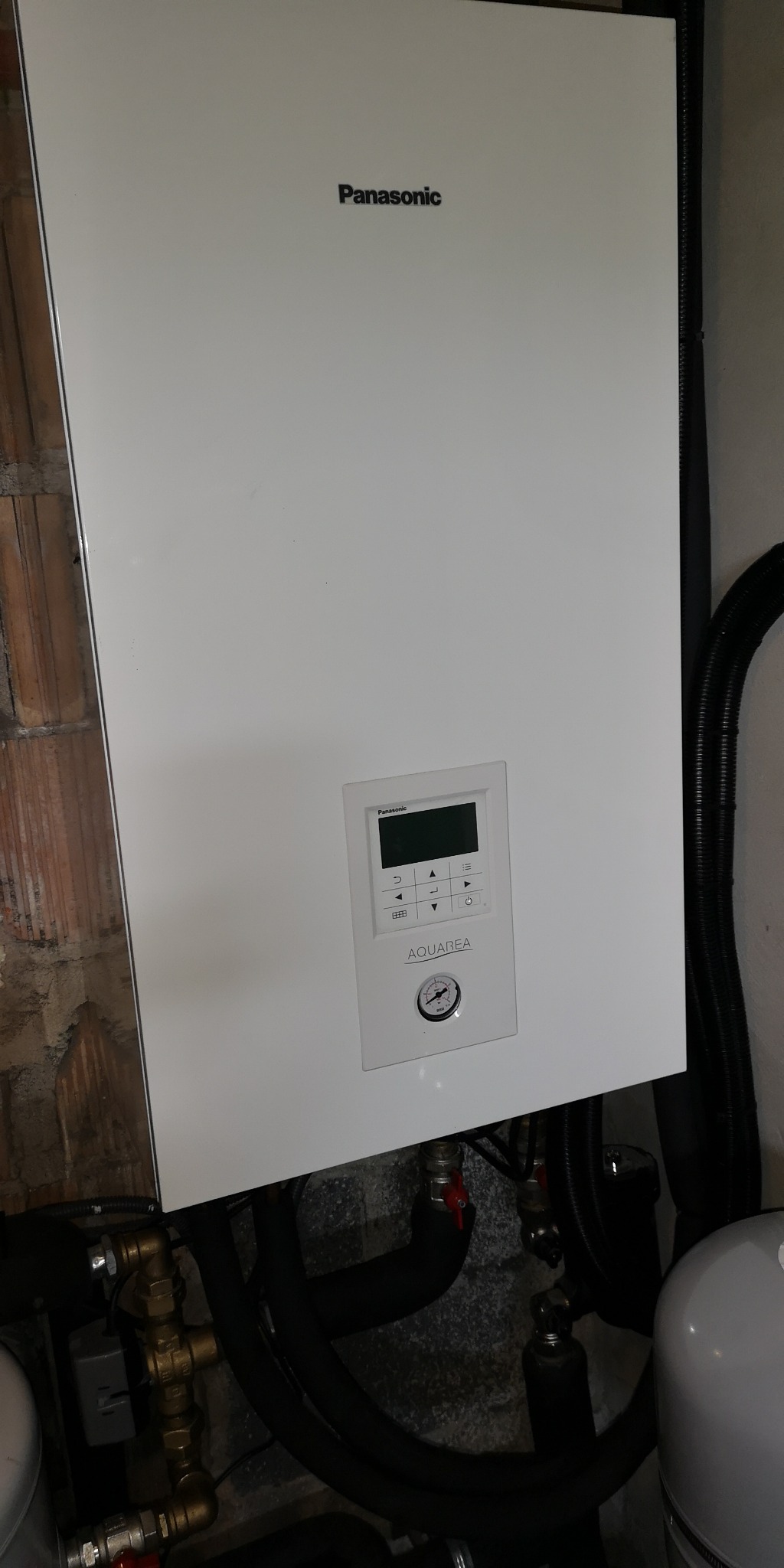 Biała jednostka wewnętrzna Panasonic Aquarea z wyświetlaczem i manometrem, zamontowana na ścianie z widocznymi rurami i zaworami.