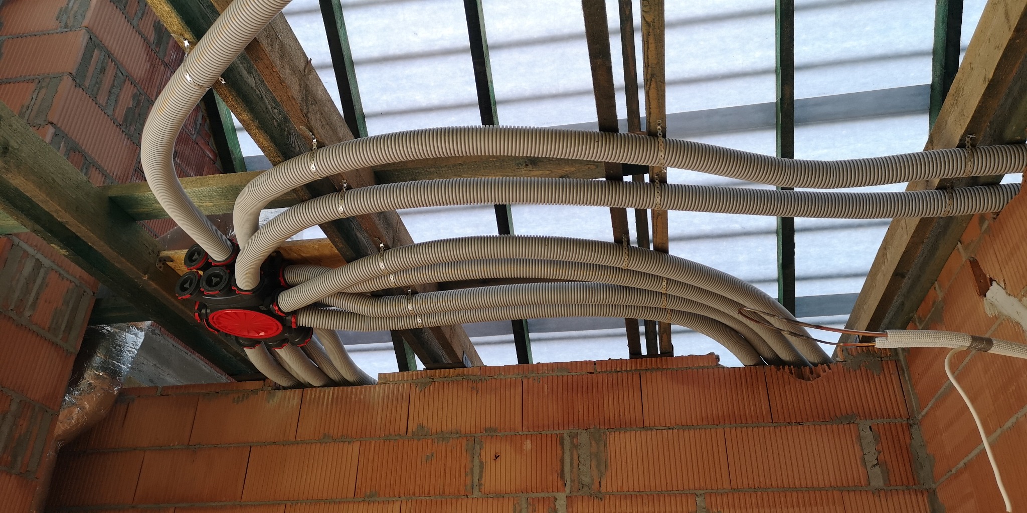 Instalacja przewodów freonowych w izolowanych rurach karbowanych, wychodzących z rozdzielacza z czerwonymi elementami, zamocowanych do drewnianej konstrukcji stropu w surowym stanie.