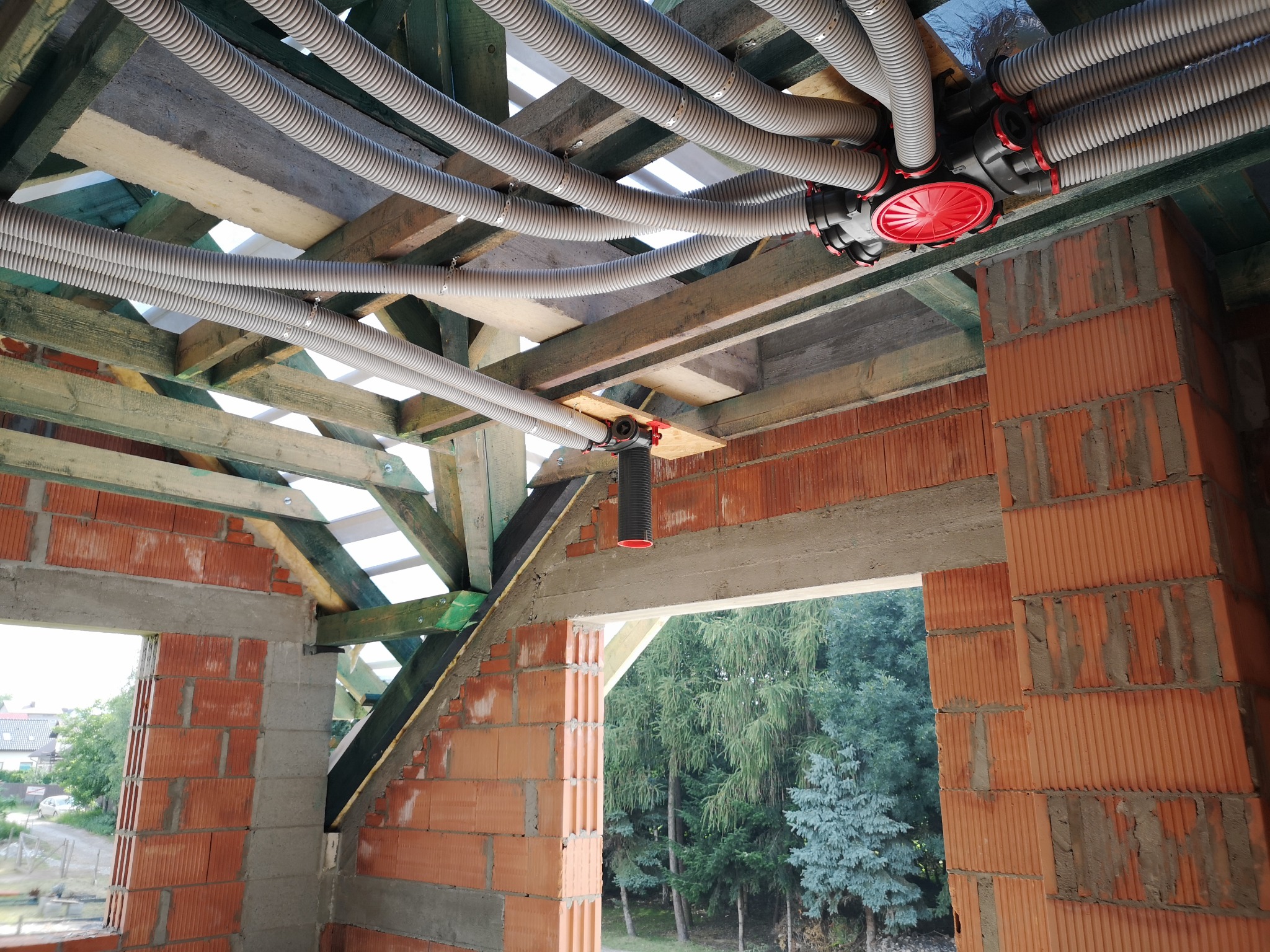 Rozprowadzenie rur wentylacyjnych rekuperacji pod drewnianą konstrukcją dachu w surowym stanie, widoczne ceglane ściany budynku w trakcie budowy.