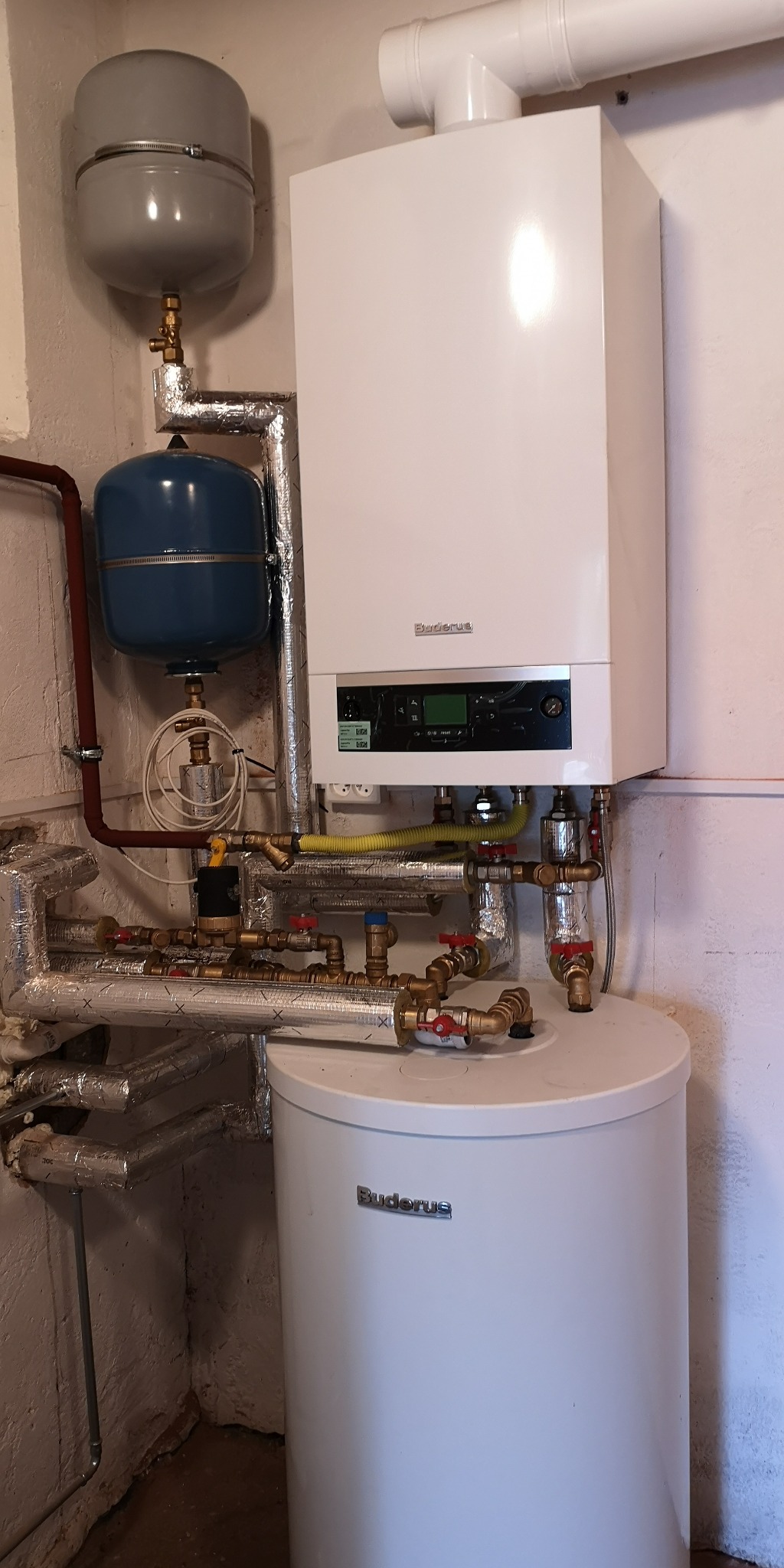 Biały piec gazowy Buderus z podłączonym zasobnikiem ciepłej wody i elementami instalacji CO, zainstalowany w piwnicy.