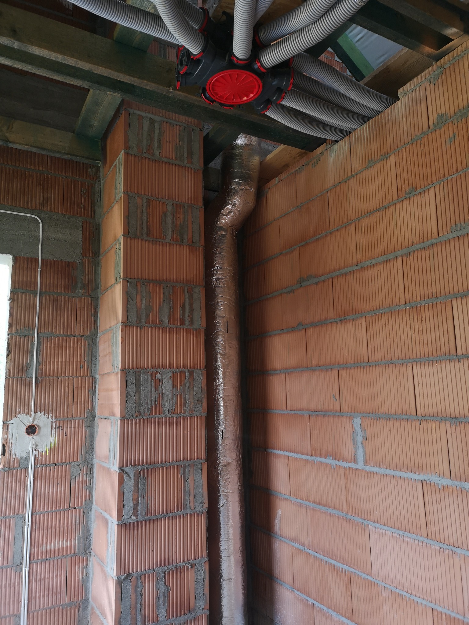 Rozprowadzenie instalacji wentylacyjnej z rekuperacją w budynku w trakcie budowy, widoczne ceglane ściany, rury wentylacyjne i rozdzielacz z czerwonymi elementami.