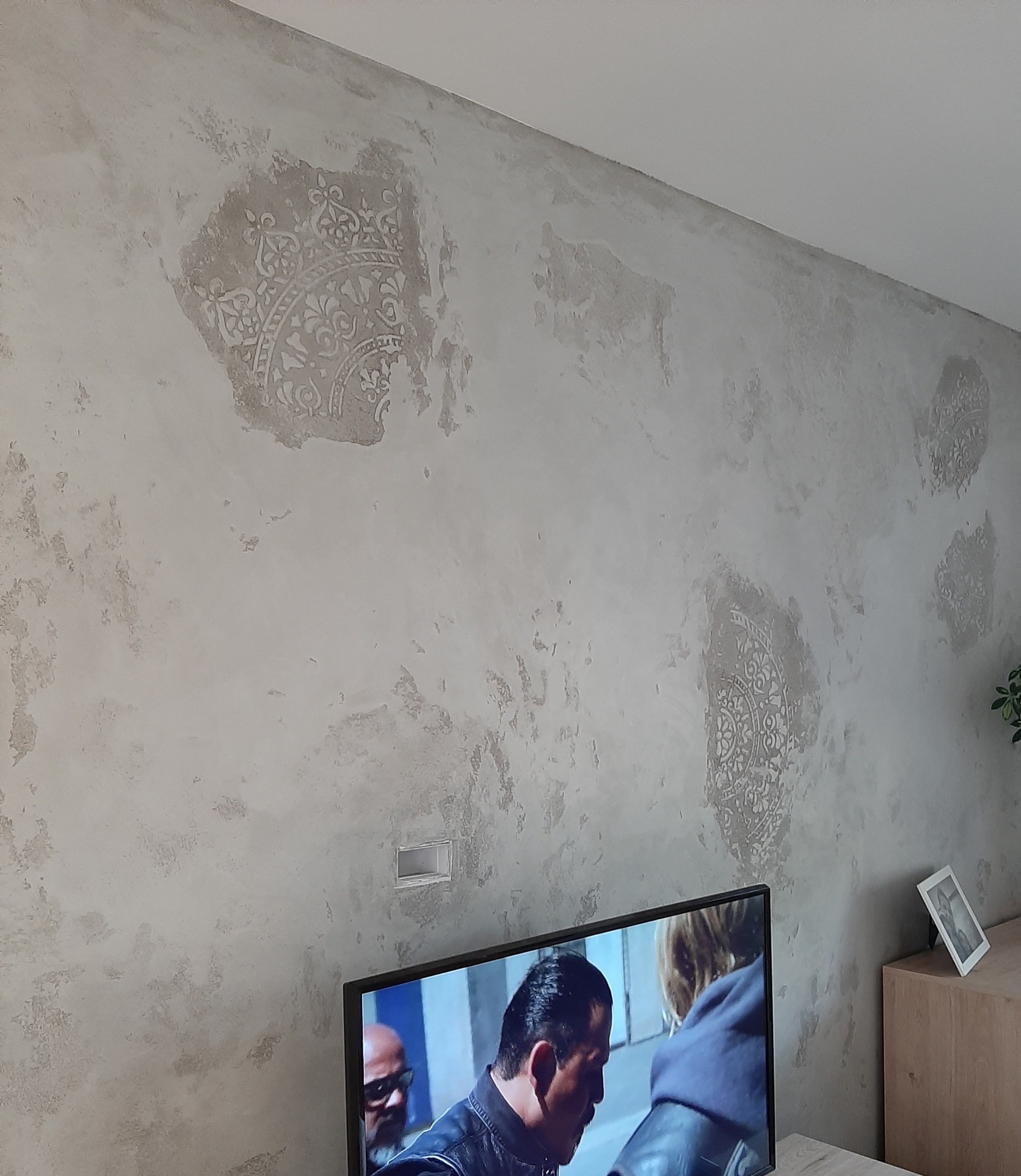 Ściana w mieszkaniu z dekoracyjną fakturą i ornamentami, widoczny fragment telewizora i mebla.
