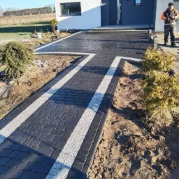 Nowoczesny podjazd z ciemnej kostki brukowej z jasnymi pasami, prowadzący do domu z szarą stolarką okienną i drzwiową, widoczny pracownik w odblaskowej kurtce.