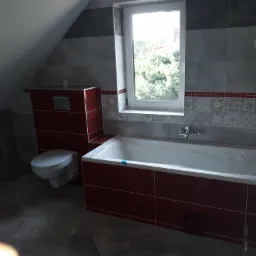 Wykończona łazienka na poddaszu z wanną w obudowie z płytek w odcieniach czerwieni, wiszącą toaletą i oknem z widokiem na zieleń.