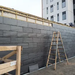 Montaż elewacji z płytek imitujących łupek, widoczna drabina i paleta z materiałem, budynek w tle.