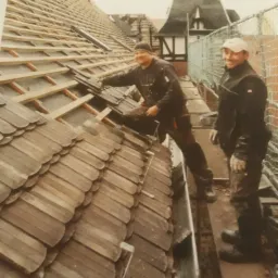 Dwóch dekarzy uśmiecha się podczas układania drewnianych dachówek na skośnym dachu budynku z wieżyczką w tle.