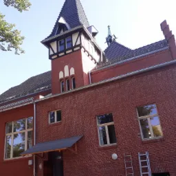 Ceglany budynek z elementami muru pruskiego i wieżyczką, widziany pod kątem z dołu, z drabinką opartą o ścianę.