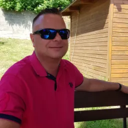 Mężczyzna w różowej koszulce polo i okularach przeciwsłonecznych siedzi na ławce przed drewnianą budką, z zielenią w tle.