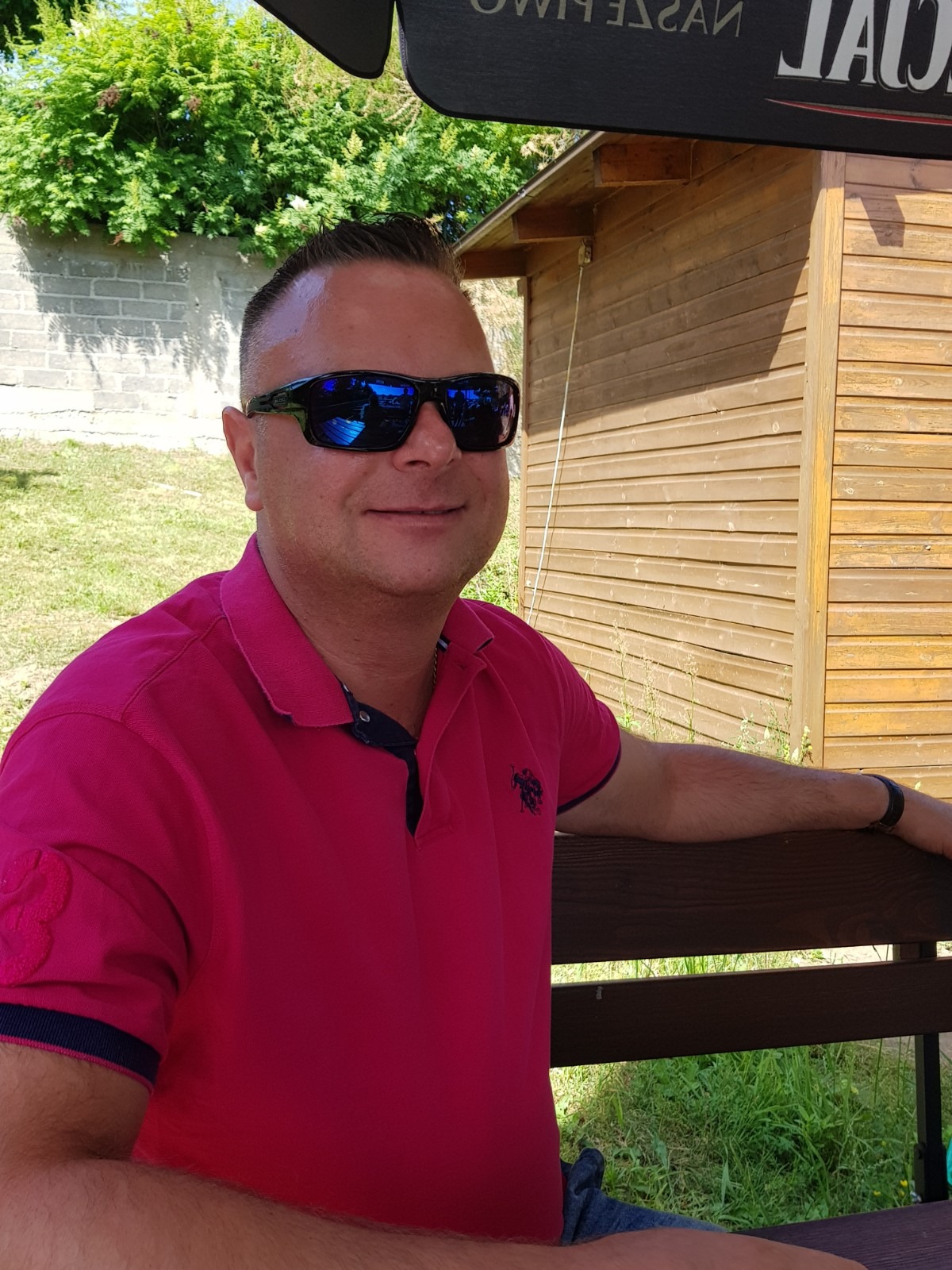 Mężczyzna w różowej koszulce polo i okularach przeciwsłonecznych siedzi na ławce przed drewnianą budką, z zielenią w tle.
