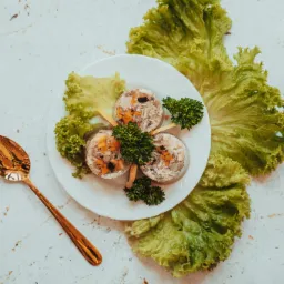 Galaretka z mięsem i warzywami na białym talerzu, udekorowana natką pietruszki i plasterkami cytryny, otoczona liśćmi sałaty, obok złota łyżka na teksturowanym tle.