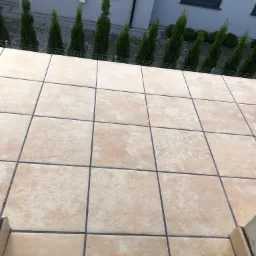 Jasne, kwadratowe płytki podłogowe na tarasie, widok z góry, z widocznymi fugami, w tle rząd zielonych tuj i fragment budynku.