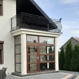 Nowoczesny dom z przeszklonym wejściem i balkonem z czarną, kutą balustradą. Widoczne brązowe ramy okien i drzwi, szara kostka brukowa oraz rząd zielonych iglaków w tle.