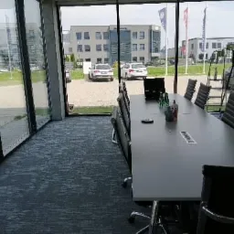 Wnętrze nowoczesnej sali konferencyjnej z długim stołem i krzesłami, widok na budynek biurowy przez duże okna, drabina oparta o ścianę w tle.