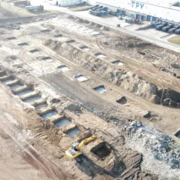 Widok z góry na plac budowy z licznymi wykopami pod fundamenty, częściowo wypełnionymi wodą, z pracującą koparką i stertami ziemi, na tle budynku magazynowego.