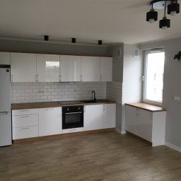 Piotr Głoćko - Jasna kuchnia z białymi szafkami, drewnianym blatem i panelami podłogowymi imitującymi drewno, oświetlona nowoczesną lampą sufitową z czarnymi kloszami.