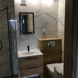 Piotr Głoćko - Nowoczesna łazienka po remoncie w Stargardzie: białe płytki imitujące marmur z szarymi żyłami, drewnopodobna szafka pod umywalką z czarną armaturą, wisząca toaleta z obudową z drewnopodobnych paneli.