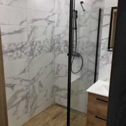 Piotr Głoćko - Nowoczesna kabina prysznicowa z czarną armaturą i szklaną ścianką, wyłożona białymi płytkami imitującymi marmur z dekoracyjnym pasem geometrycznym, drewnopodobna podłoga i szafka pod umywalkę...