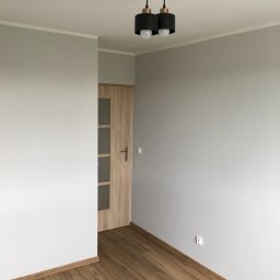 Piotr Głoćko - Pomalowane na szaro ściany w pustym pokoju z drewnianą podłogą i drzwiami, widoczny nowoczesny czarny żyrandol z dwoma kulistymi lampami.