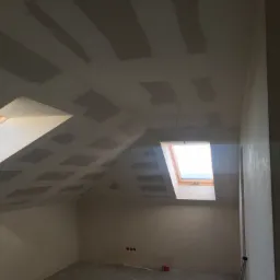 Poddasze w trakcie prac wykończeniowych: widoczne łączenia płyt gipsowo-kartonowych na skośnym suficie, przygotowane do dalszej obr&oacute;bki, z dwoma oknami dachowymi w drewnianych ramach i surowymi...