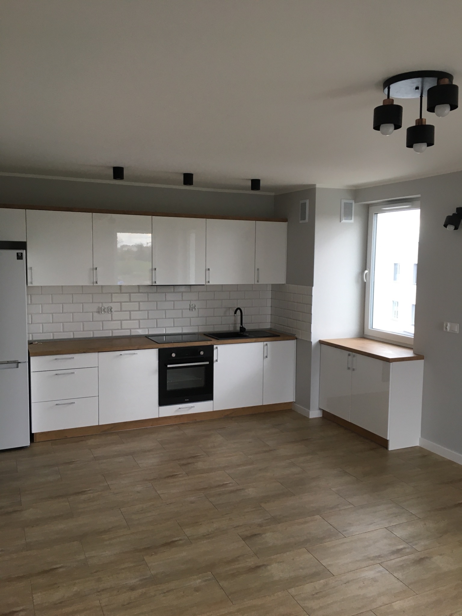 Jasna kuchnia z białymi szafkami, drewnianym blatem i panelami podłogowymi imitującymi drewno, oświetlona nowoczesną lampą sufitową z czarnymi kloszami.