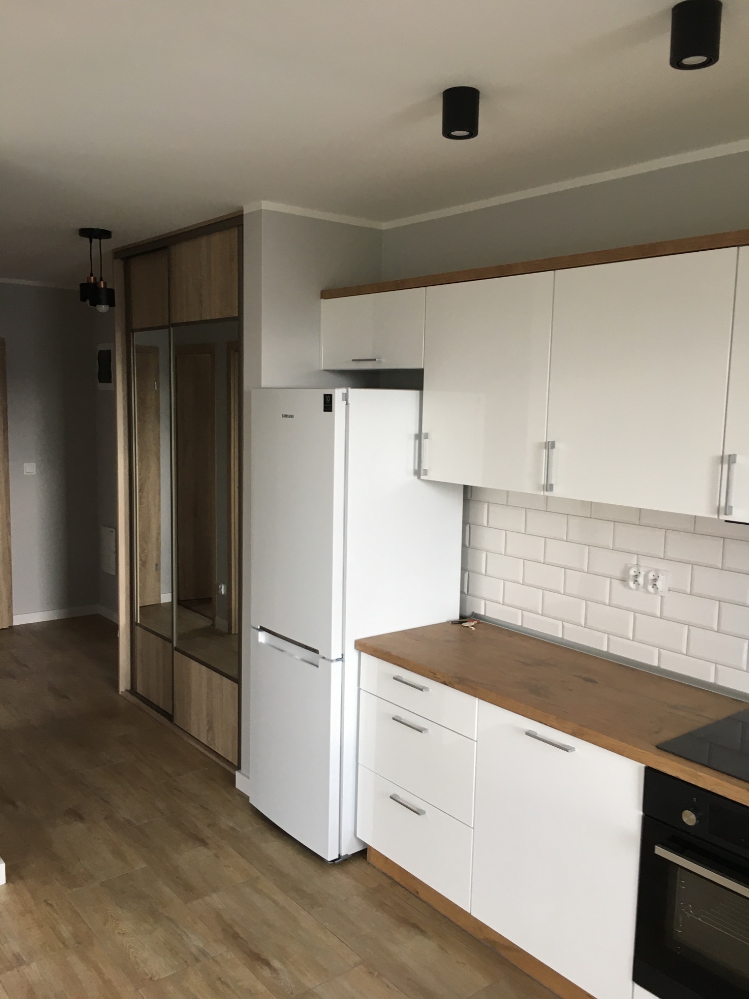 Nowoczesna kuchnia z białymi szafkami, drewnianym blatem i płytkami typu metro na ścianie, obok szafa wnękowa z lustrzanymi drzwiami i oświetleniem punktowym.