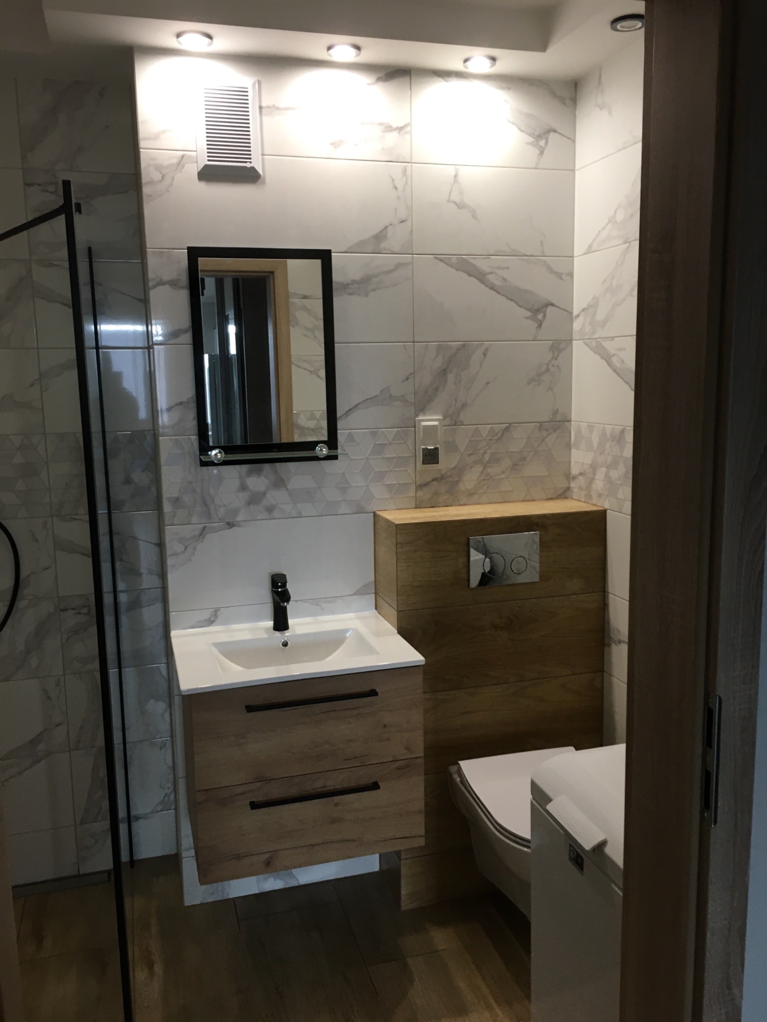 Nowoczesna łazienka po remoncie w Stargardzie: białe płytki imitujące marmur z szarymi żyłami, drewnopodobna szafka pod umywalką z czarną armaturą, wisząca toaleta z obudową z drewnopodobnych paneli.