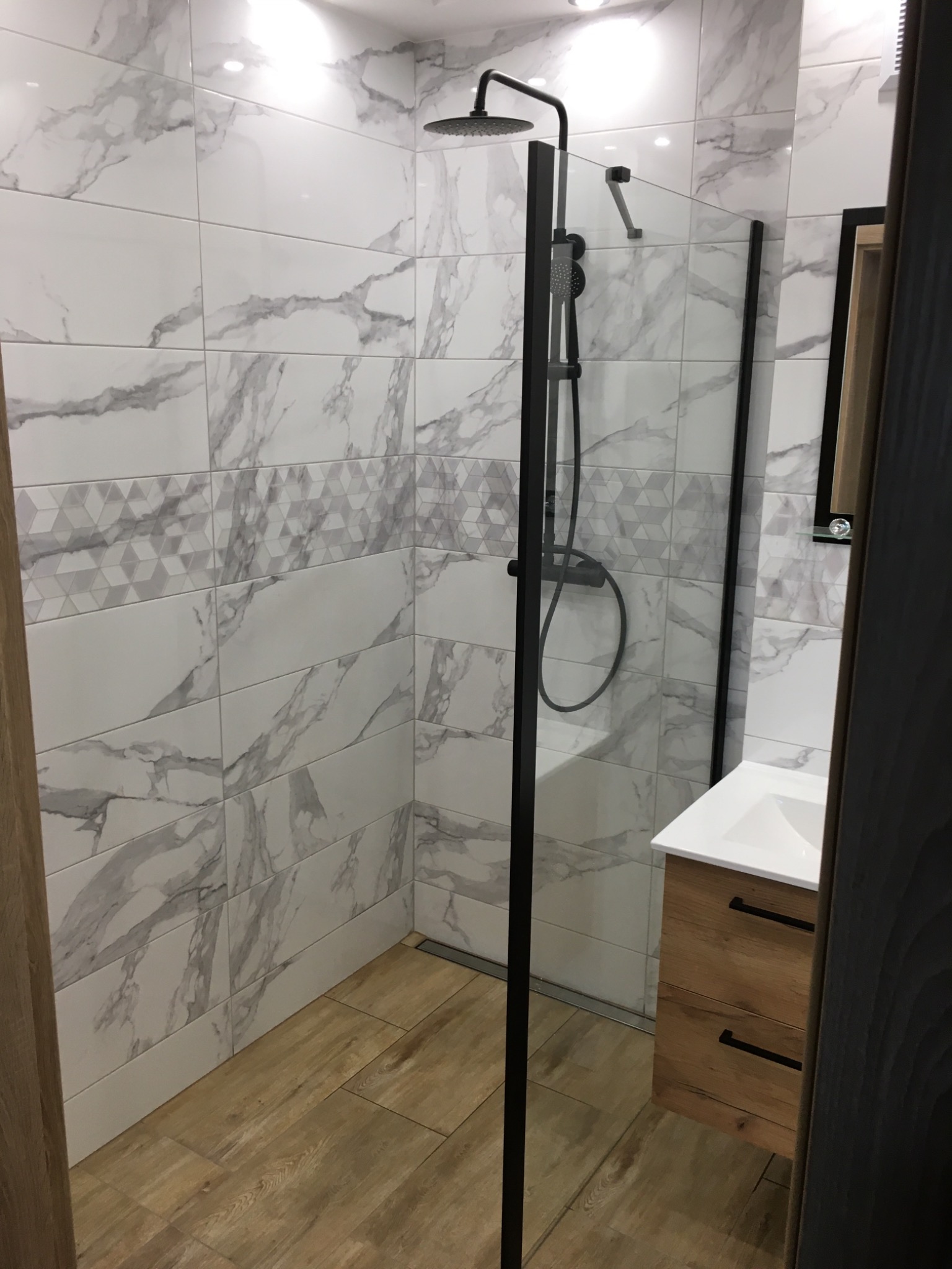 Nowoczesna kabina prysznicowa z czarną armaturą i szklaną ścianką, wyłożona białymi płytkami imitującymi marmur z dekoracyjnym pasem geometrycznym, drewnopodobna podłoga i szafka pod umywalkę...