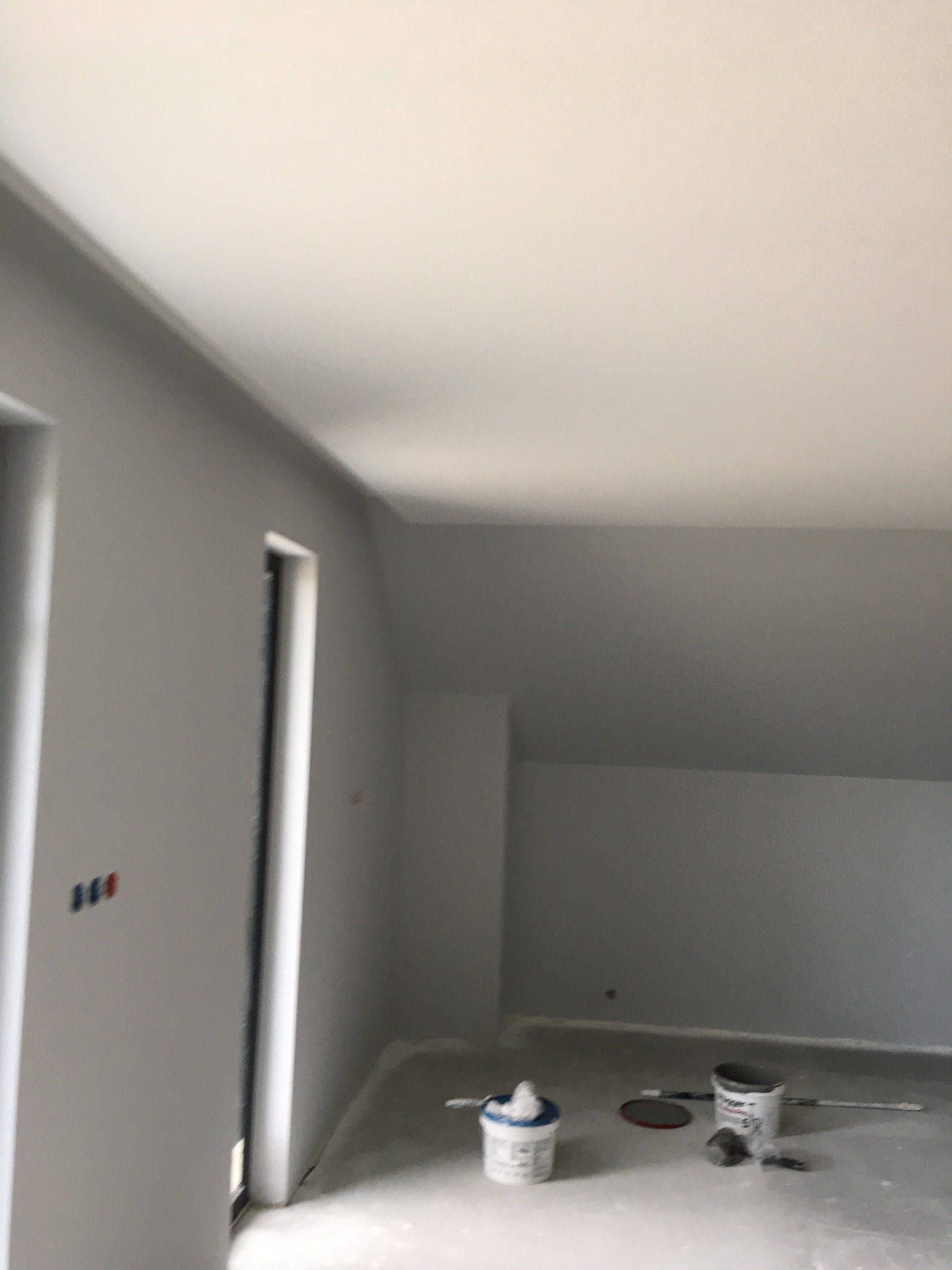 Szare wnętrze w trakcie malowania, widoczne puszki z farbą na podłodze, ściany częściowo pomalowane na szaro, widoczne gniazdka elektryczne.